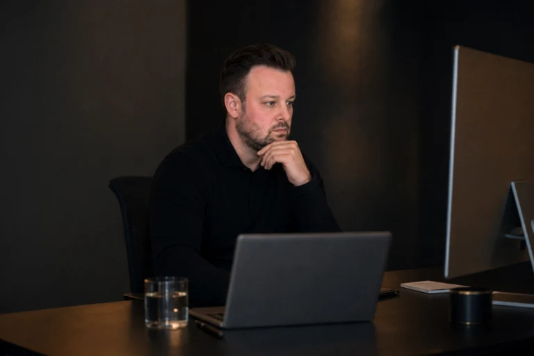 David Keiser sitzt nachdenklich vor Monitor und Laptop in einem dunklen Büro